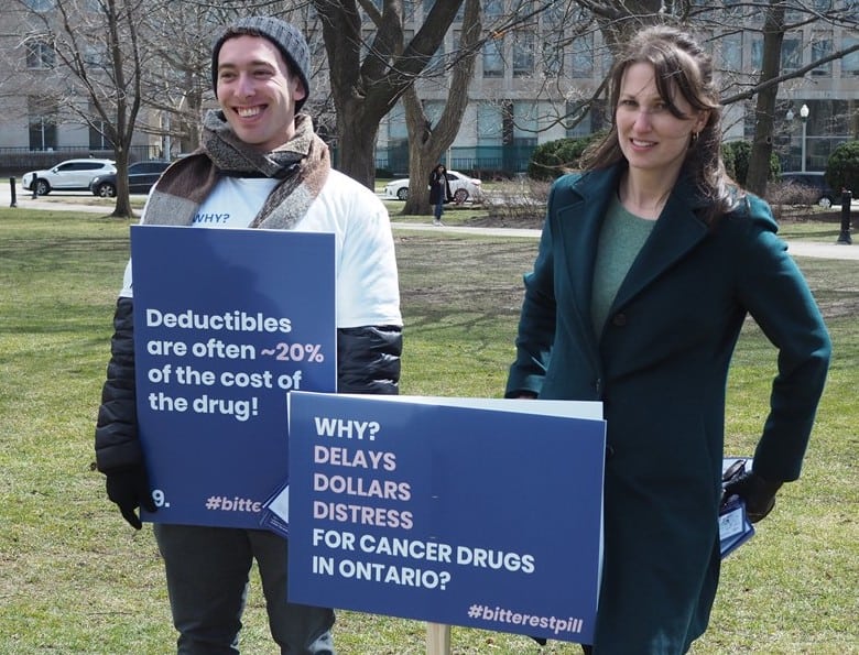 Two people standing on the lawn of the Ontario legislature; one holds a sign saying “Deductibles are often about 20% of the cost of the drug #BitterestPill”, and the other holds a sign saying “Why? Delays, dollars, distress for cancer drugs in Ontario? #BitterestPill”.